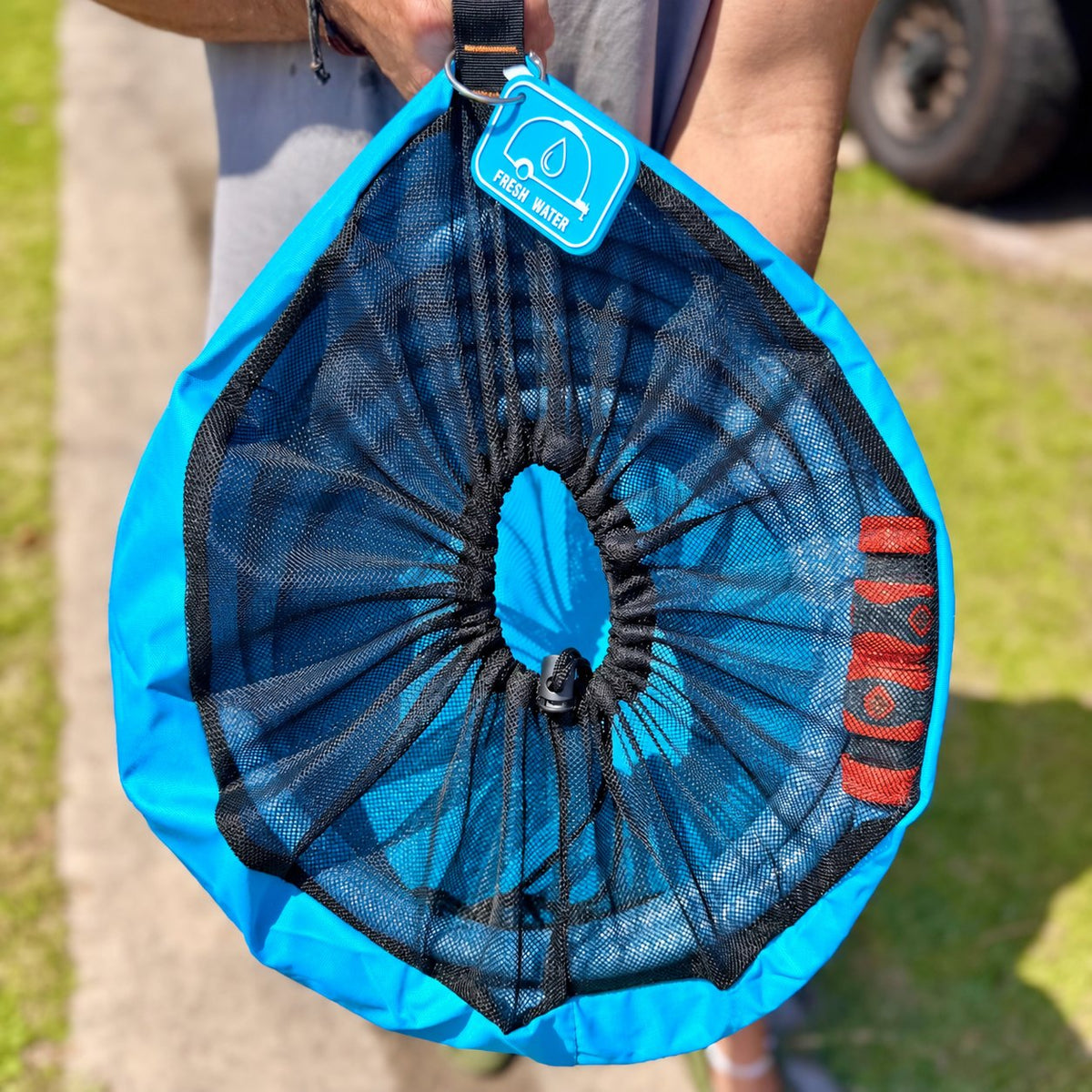 Blue drinking water hose bag held by its handle, showing the mesh drawstring top and rubber tag on a metal ring